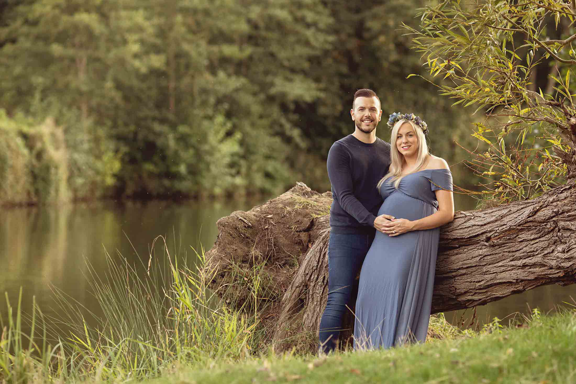 Couple sitting on large fallen tree next to a lake. Pregnant lady holding her baby bump by Maternity Photographer in Medway, Kent