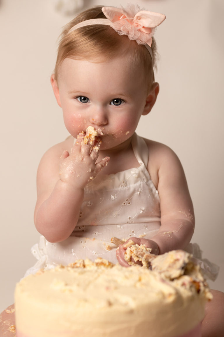 Cake Smash for babies first birthday. Baby girl wearing white romper and pink headband eating and smashing cake.