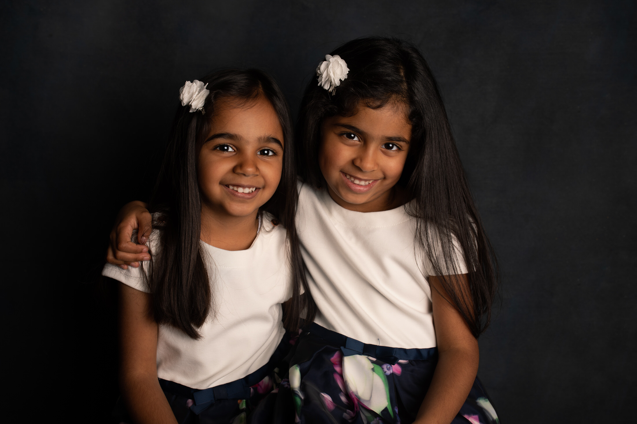 Children photoshoot - siblings wearing matching dresses cuddling on dark backdrop