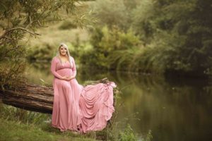 Maternity photoshoot with mum wearing pink dress sitting next to lake