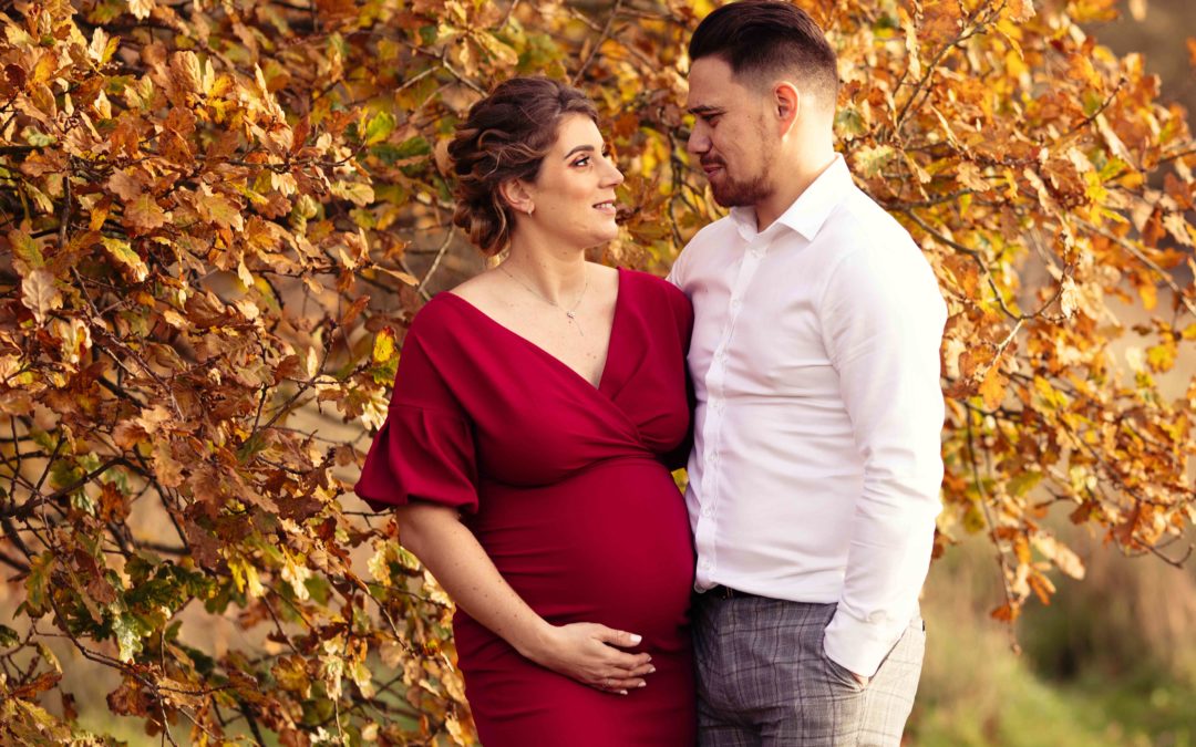 Maternity photoshoot - Pregnant lady with husband in Medway Kent. Posing in front of a tree wearing red dress holding baby bump.