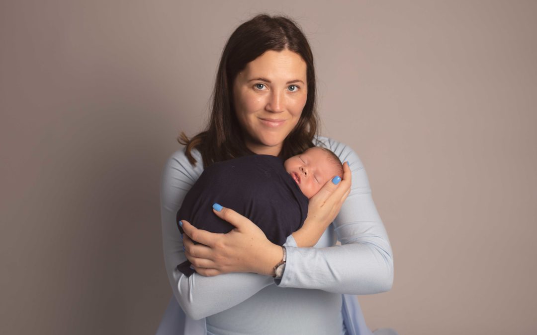 New mum holding her newborn son - Mum is wearing light blue dress with newborn wrapped in a navy wrap.. Photographed in Medway, Kent