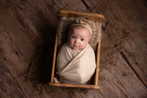 Newborn Baby Girl photo - baby is swaddled in a cream wrap in a wooden bed. Newborn girl is wearing a headband. Photographed in Medway, Kent.