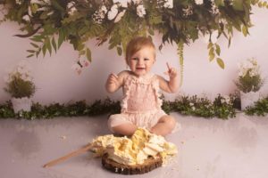 Cake Smash photo of One year old girl in pink outfit. There is a smashed cake and wooden spoon and she is cheering.