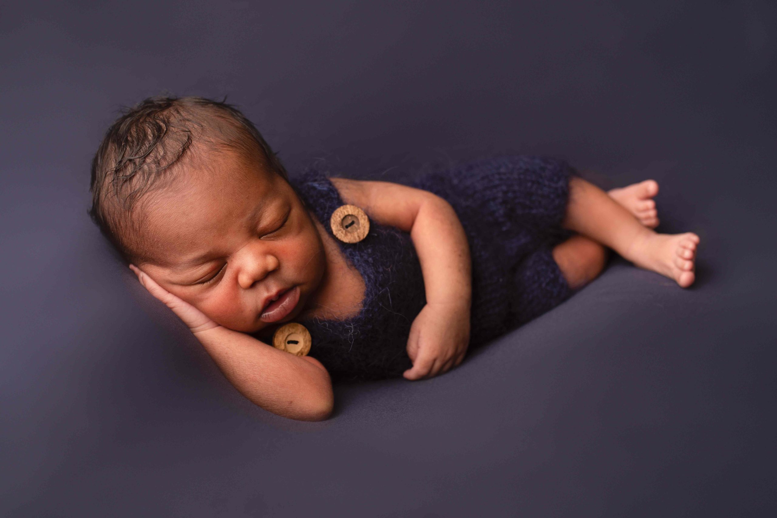 newborn boy laying on his side in a navy romper with head on one hand by Newborn Photographer Medway