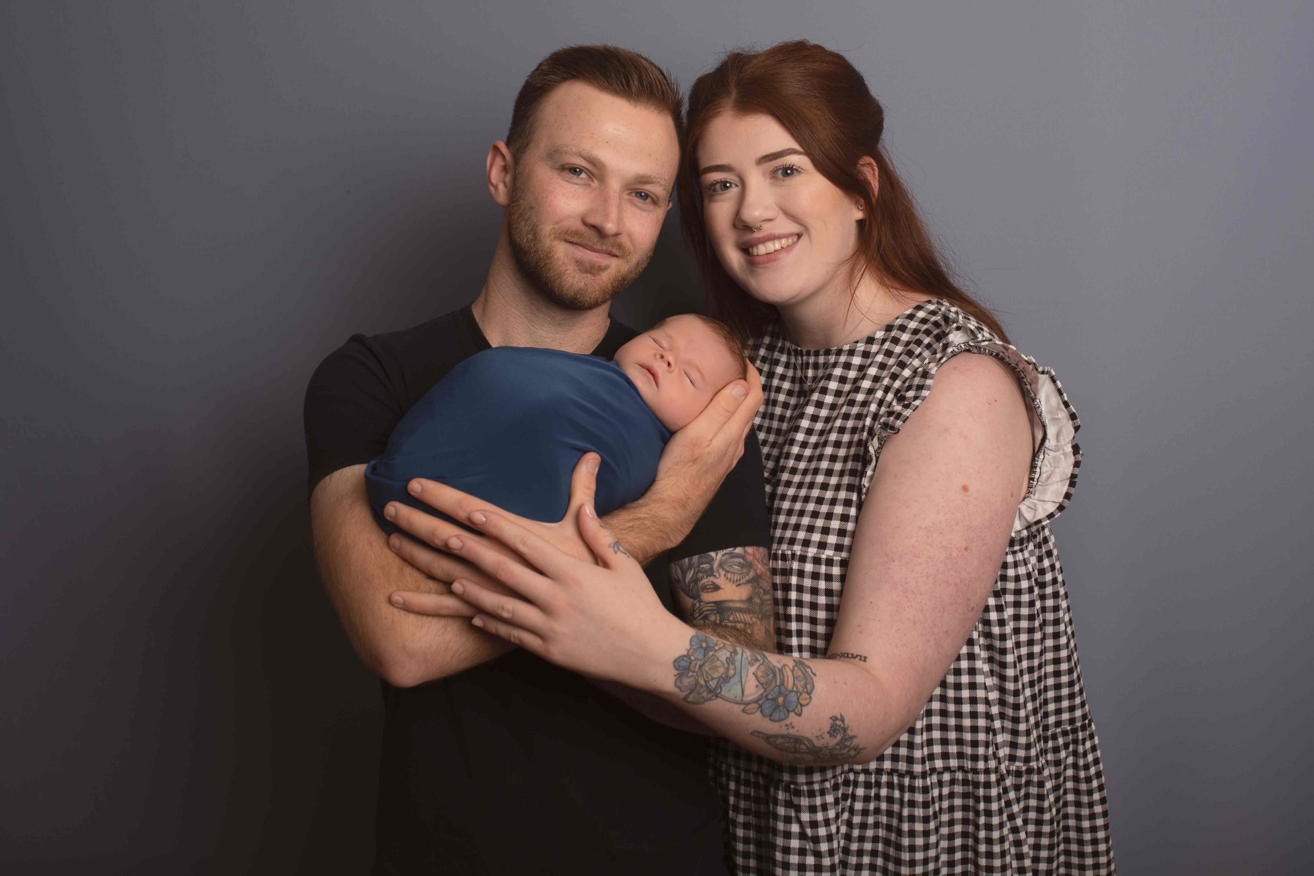 Dad holding baby boy who is wrapped in blue wrap with mum cuddling into them by Newborn Photographer Medway