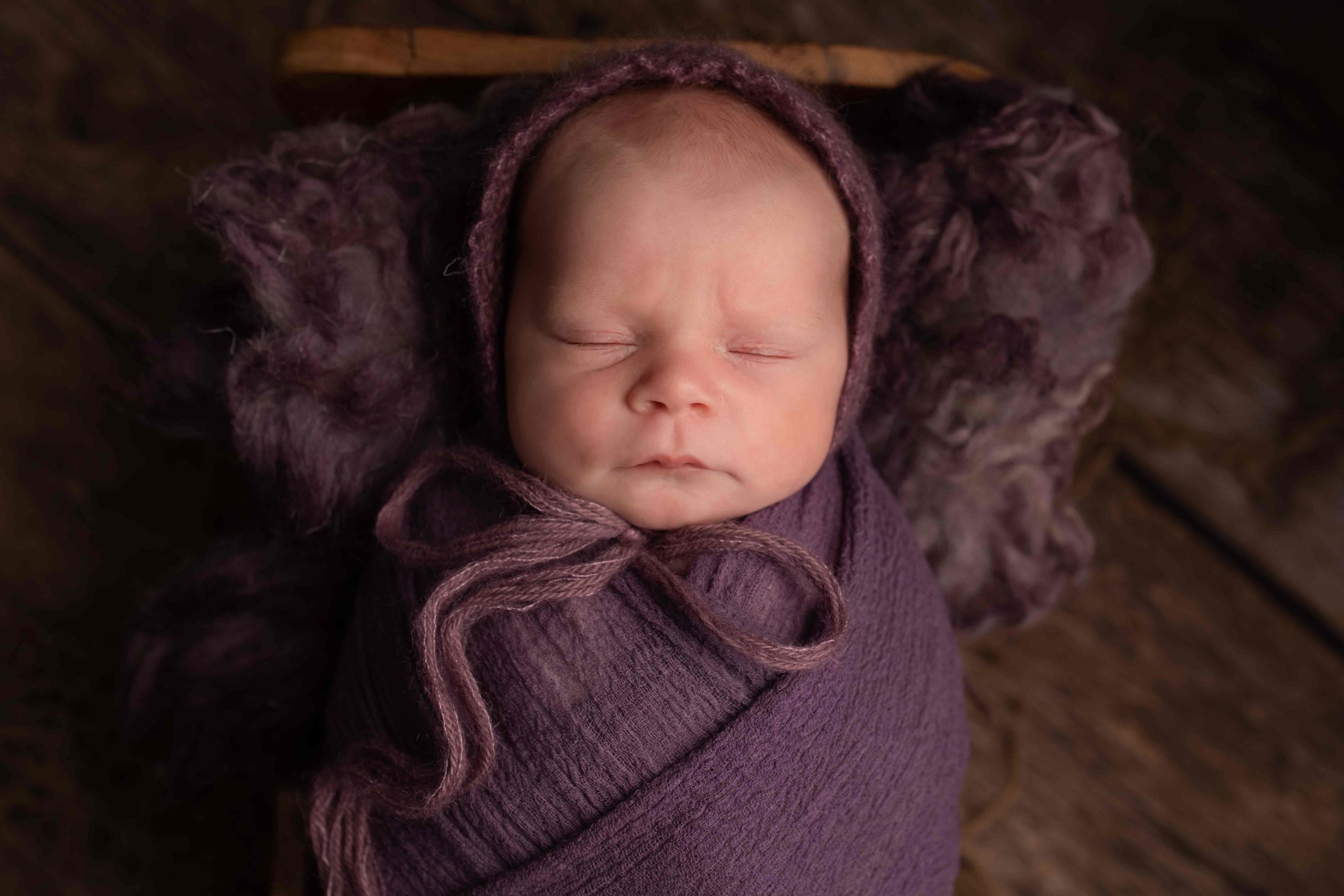 Newborn baby girl wrapped in purple wrap laying in wooden bed by Newborn Photographer in Medway