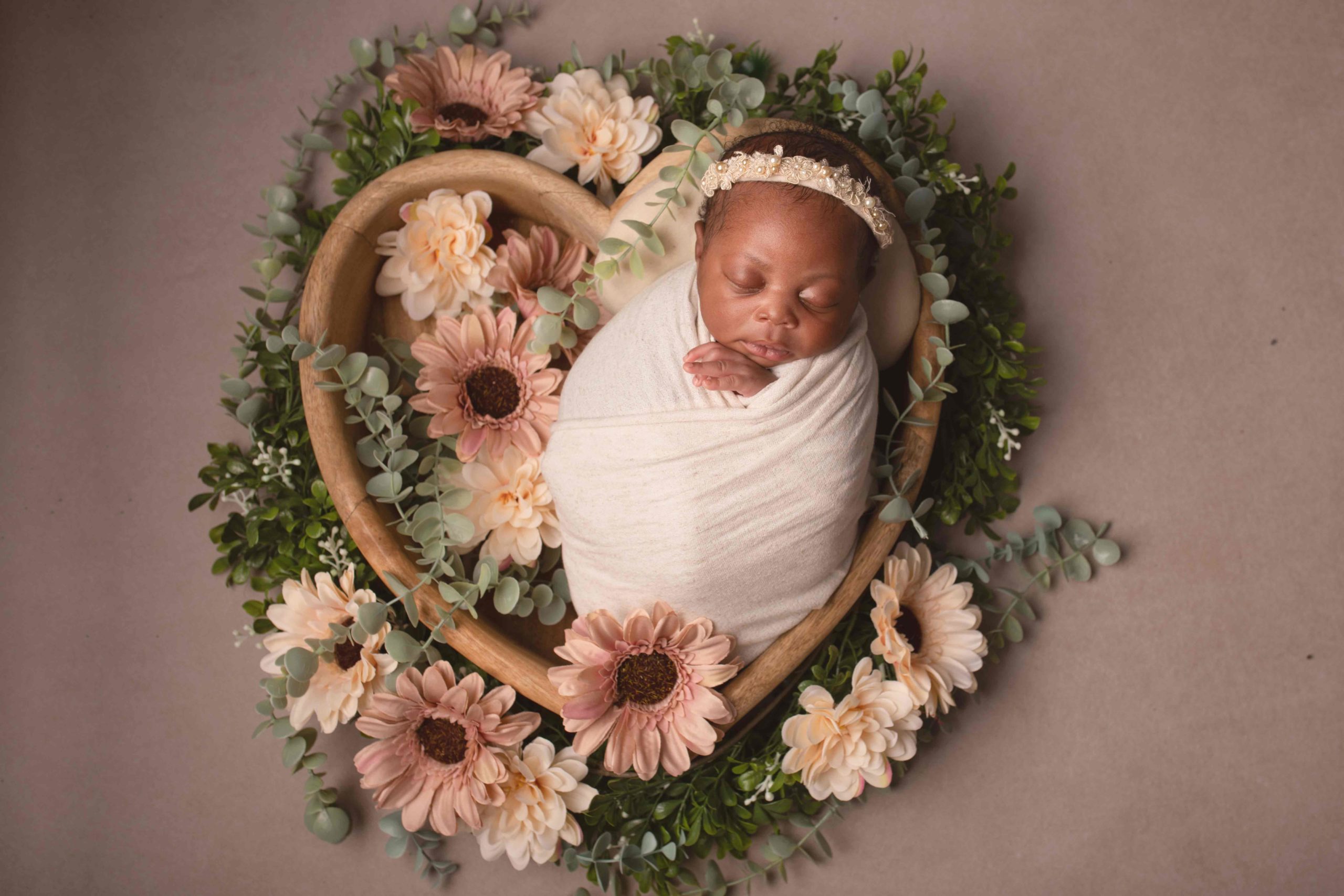 Baby girl in white wrap laying in hear shaped bowl surrounded by flowers by Newborn Photographer in Medway