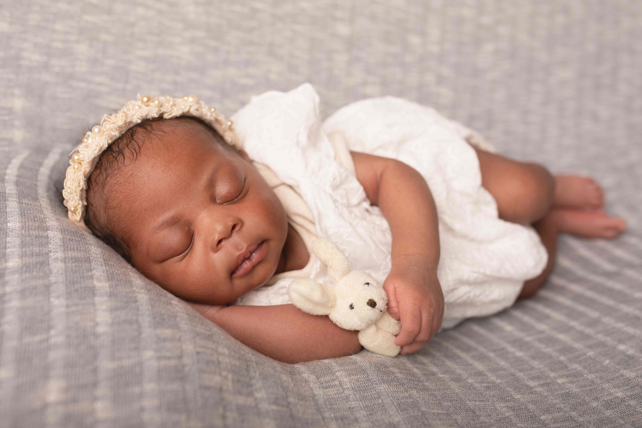 newborn girl wearing white outfit laying on grey backdrop cuddling teddy by Newborn Photographer in Medway