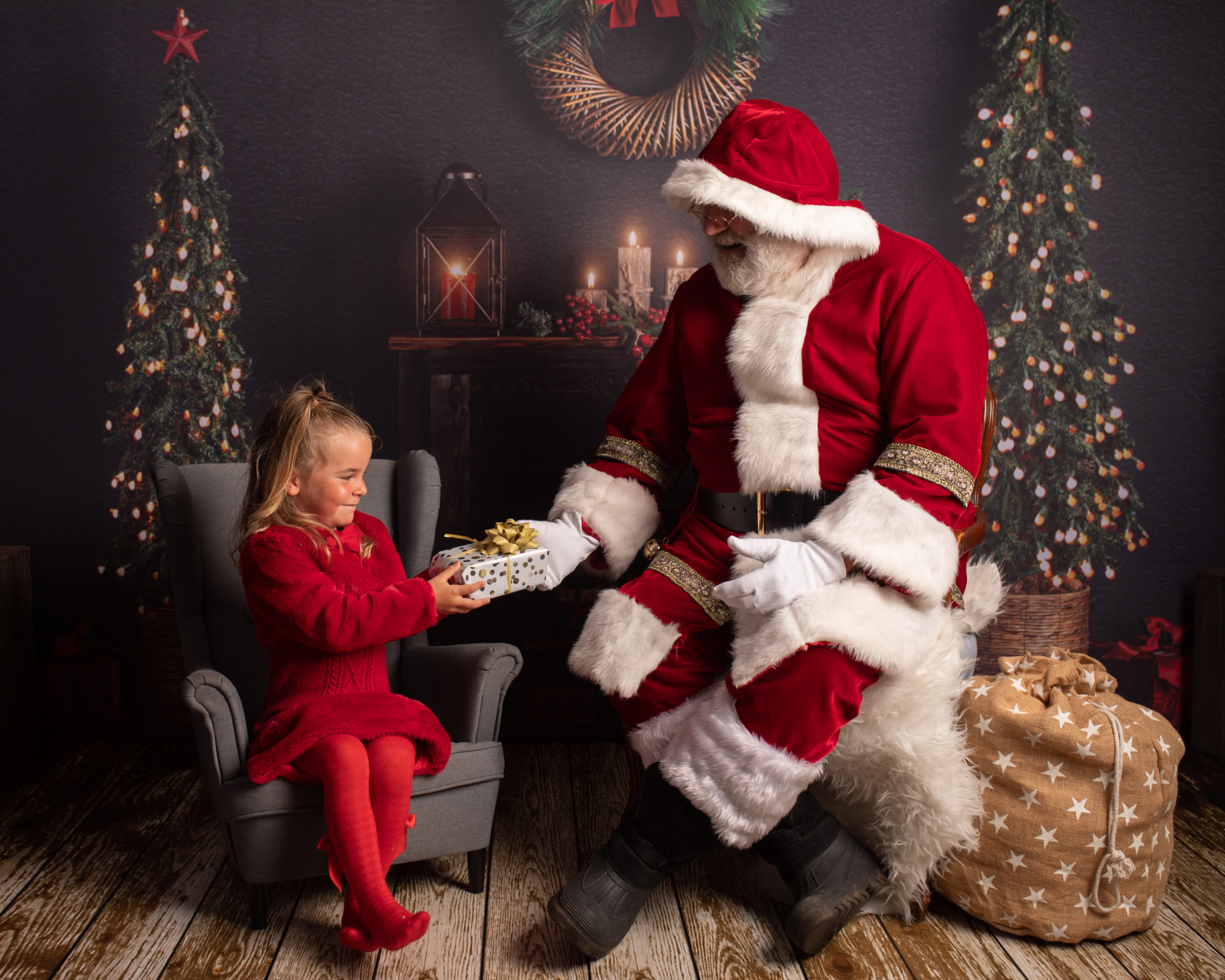 Santa handing little girl a present in front of fireplace and Christmas trees. Photographed by Kent Family Photographer