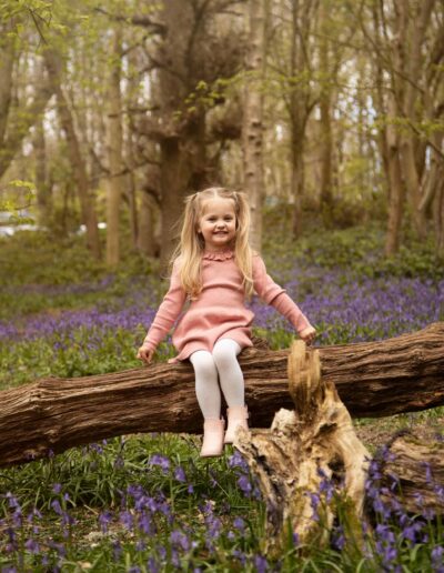 Little girl sat on fallen tree branch amongst bluebells. Photographed by Hannah Cornford who is a Family photographer based in Medway, Kent