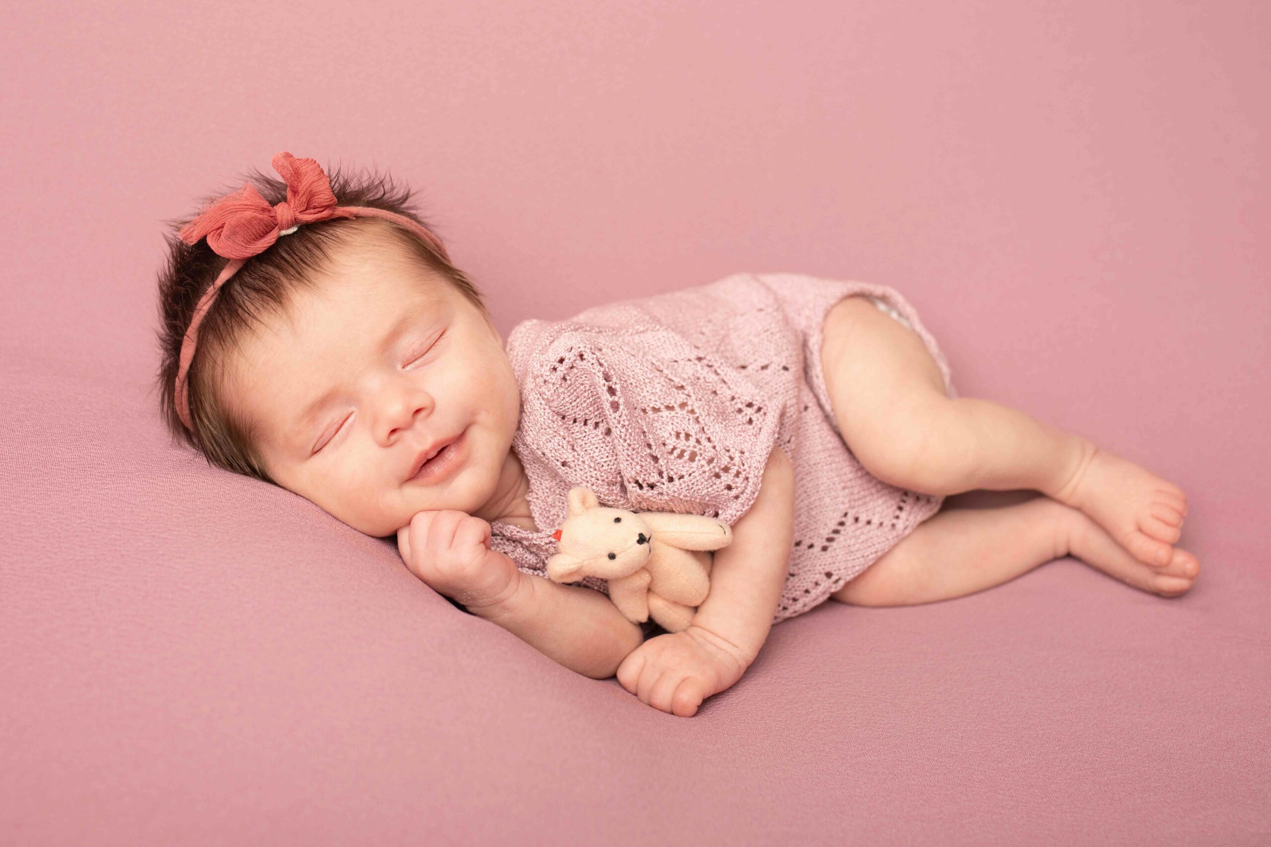 Newborn baby girl in pink laying on bed cuddling teddy. New parents to be photographed in my Photography studio in Medway Kent.