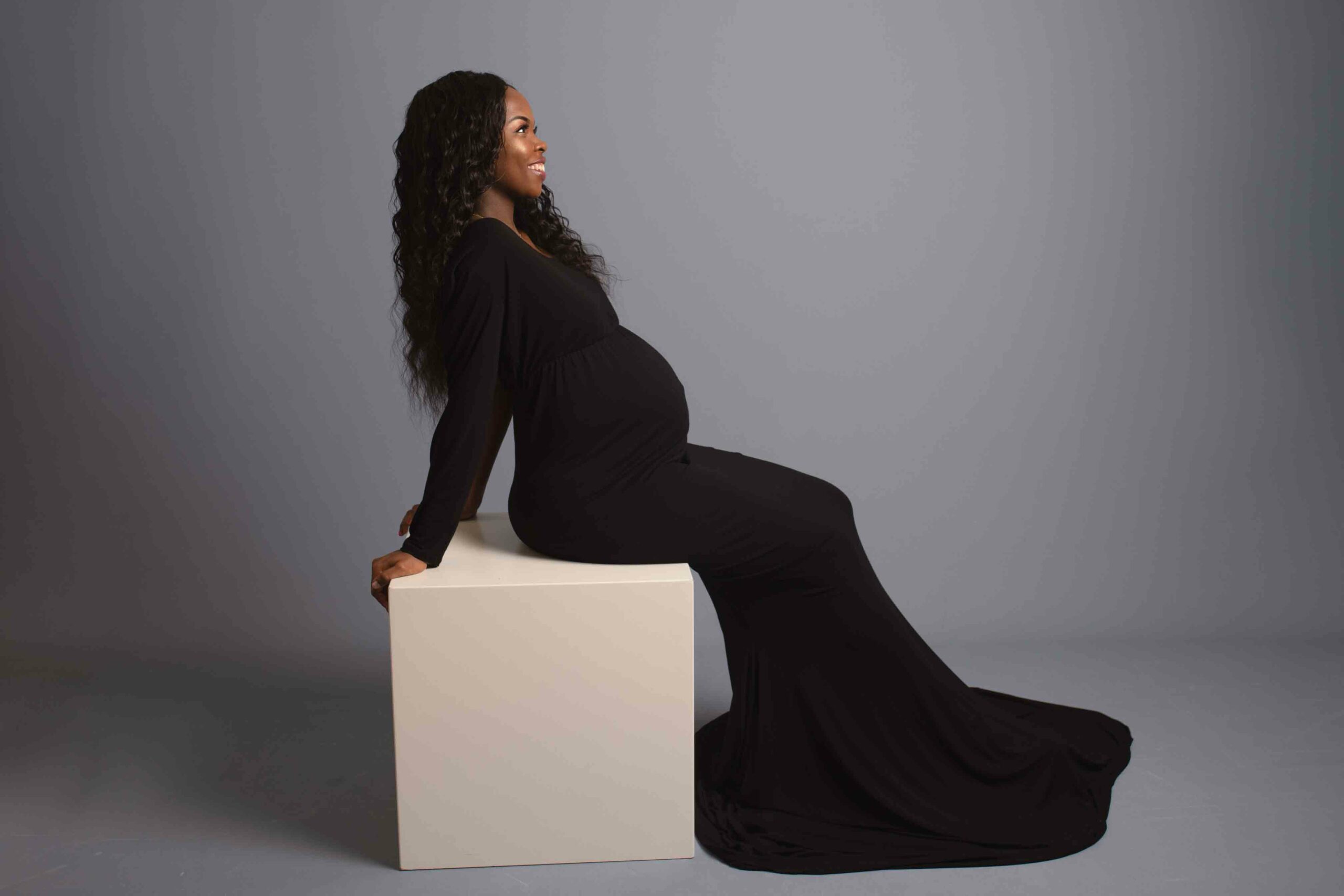 Pregnant mum to be leaning on white block wearing maternity black dress