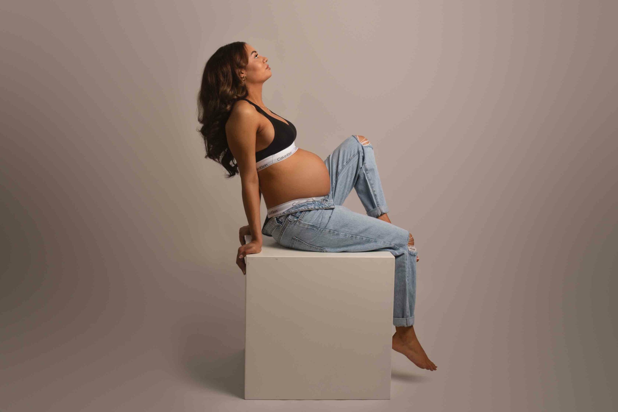 Pregnant mum photographed in jeans and black sports bra. Sitting on white block with one knee up. Photographed in my studio in Strood, Kent