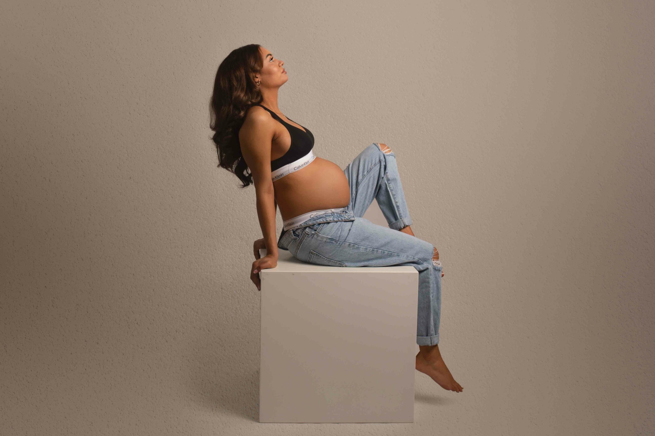 Pregnant mum photographed her maternity photos in jeans and black sports bra. Sitting on white block with one knee up. Photographed in my studio in Strood, Kent