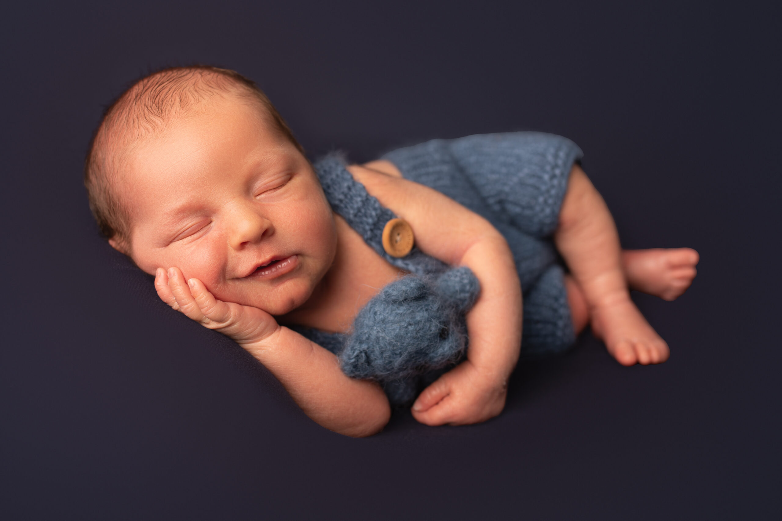 Newborn baby boy laying on his side cuddling a blue teddy bear. Photographed in Hannah Cornford studio in Medway, Kent.