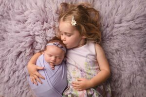 A baby girl wrapped in a lilac wrap being held by big sister. A beautiful sibling photo Photographed in Hannah Cornford Photography studio in Medway Kent. 
