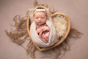 Newborn baby girl photographed in a wooden heart bowl wrapped in a cream wrap. Photographed in Medway, Kent by Hannah Cornford Photography