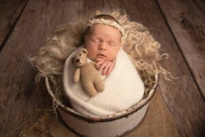Newborn baby girl wrapped in a cream wool wrap holding a teddy bear. She is wearing a beautiful cream headband and resting in a cream bucket. Photographed in Hannah Cornford Photography studio in Medway Kent. 