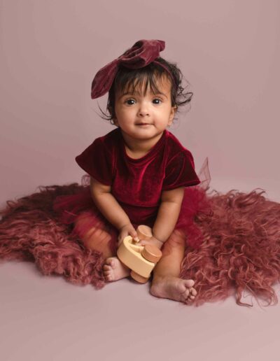 Baby girl wearing burgundy red tutu dress sitting on pink background with matching hair bow. Photographed by Hannah Cornford in Medway, Kent