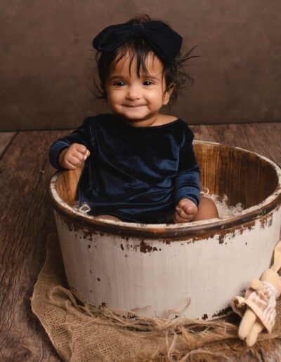 Baby girl wearing navy blue outfit. She is sitting in a bucket. Taken by Hannah Cornford Photography a Family and Newborn photographer based in Medway, Kent.
