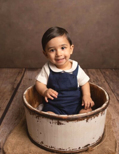 Baby boy sitting in a wooden bucket. He is wearing a navy blue dungaree set with brown hair and eyes. Taken by Hannah Cornford Photography a Family and Newborn photographer based in Medway, Kent.