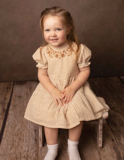 A little girl sitting on a wooden stall wearing a beige dress with long blonde hair. Taken by Hannah Cornford Photography a Family and Newborn photographer based in Medway, Kent.
