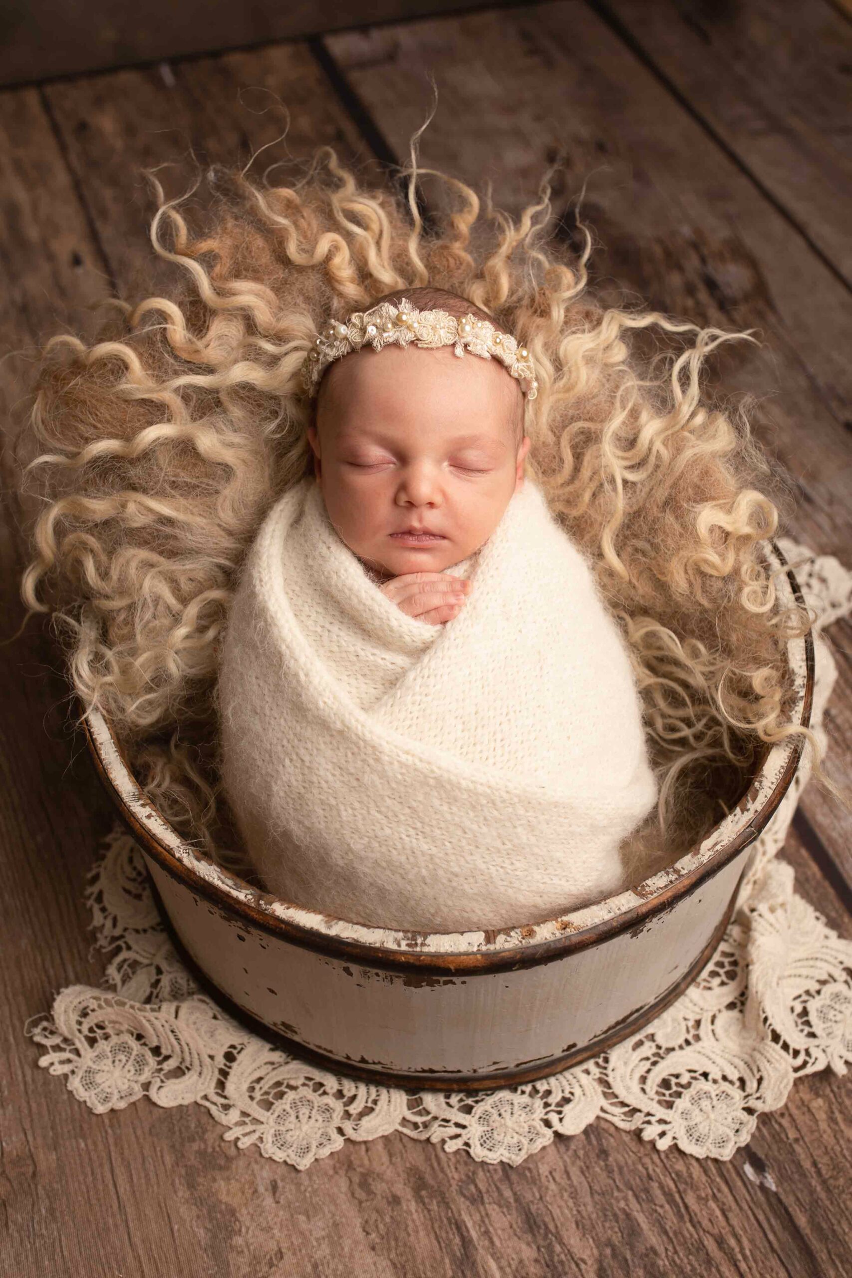 Newborn baby girl wrapped in cream soft wrap. Wearing a pearl headband in a cream wooden prop. Photographed in Medway Kent by Hannah Cornford 