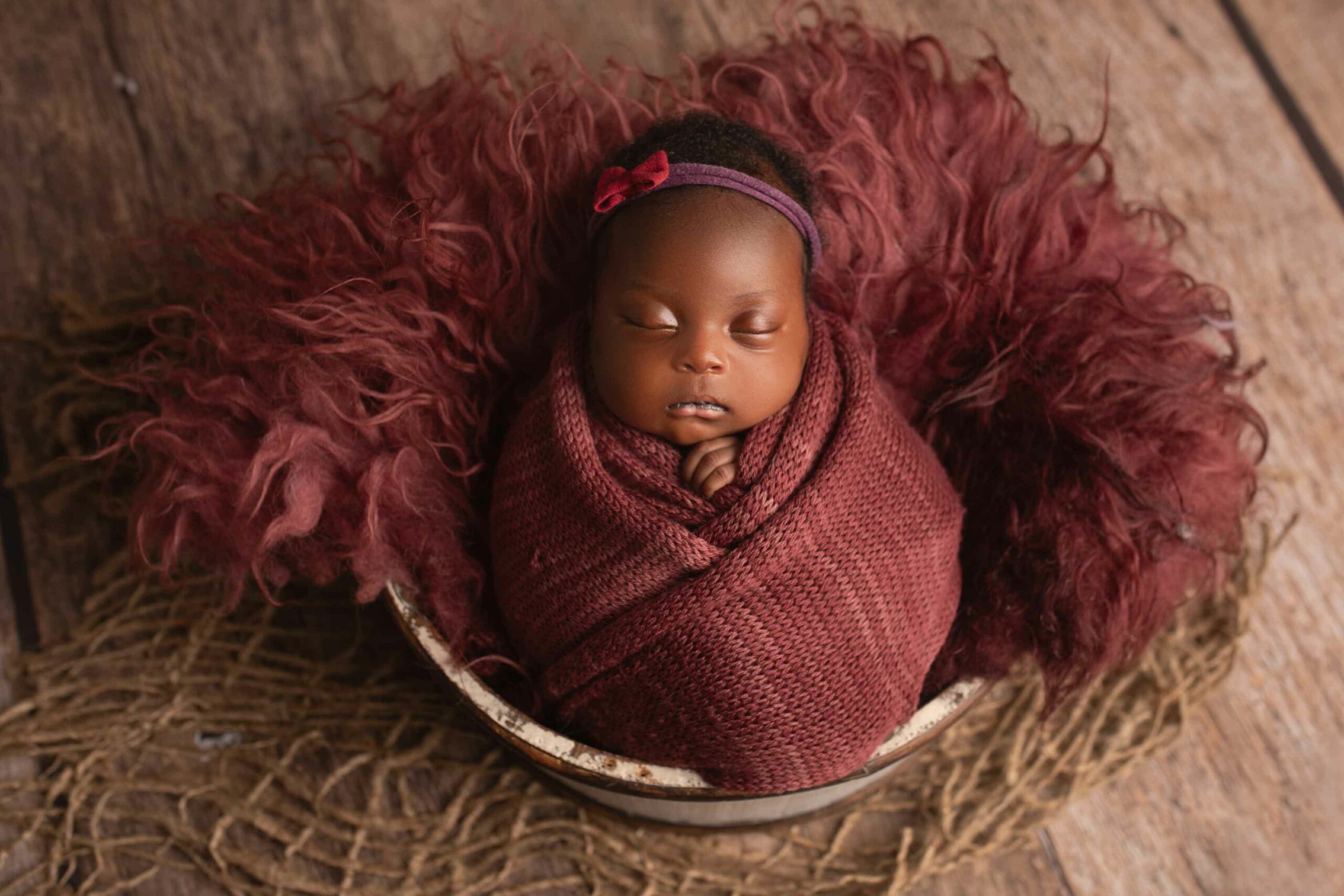 Beautiful baby girl wrapped in burgundy wrap laying on red fur. Propped up in wooden bucket. Photographed in Medway by Hannah Cornford Photography